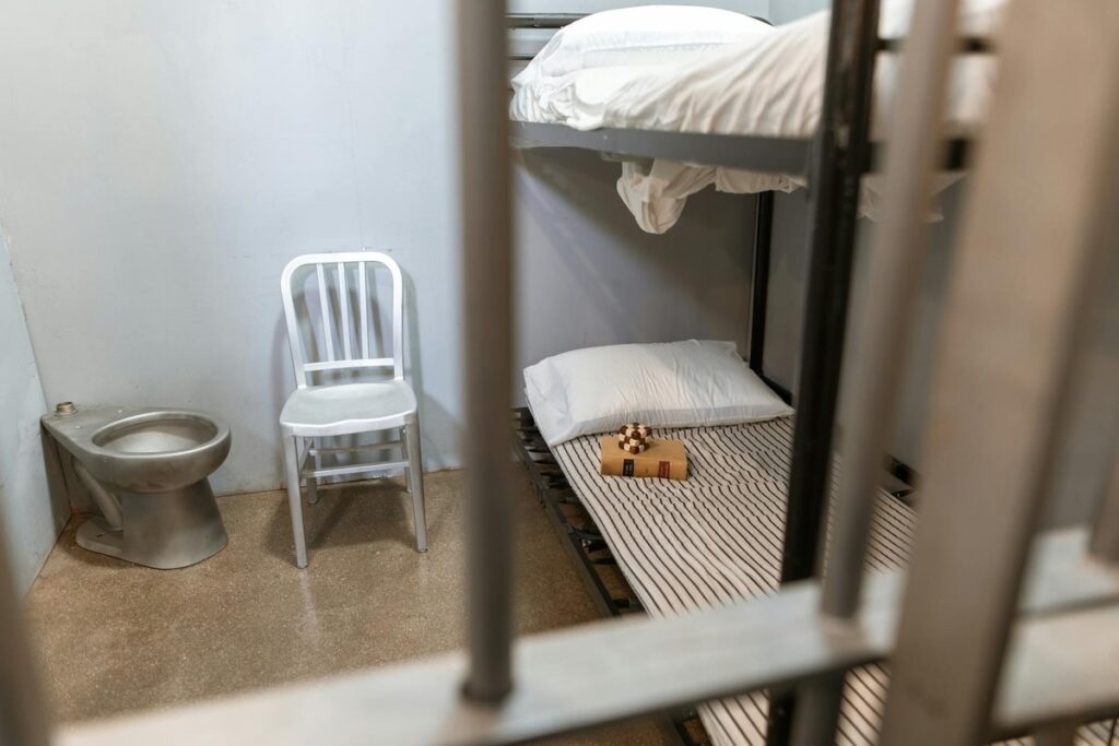 Detailed view of a clean prison cell featuring a bunk bed, metal chair, and toilet.