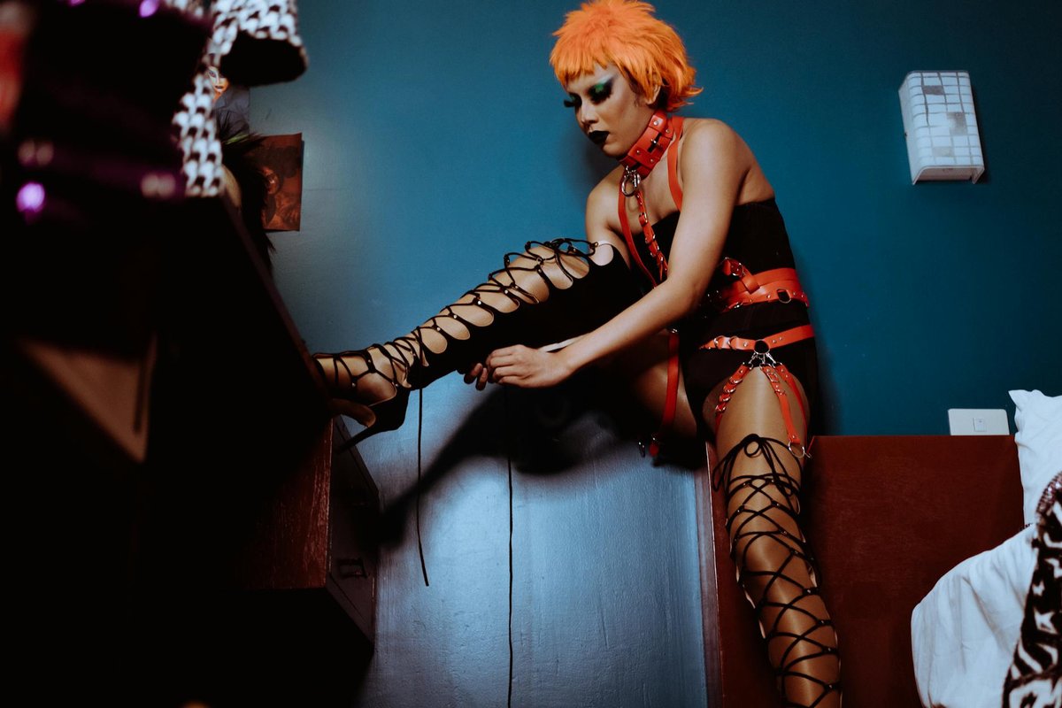 Low angle of ethnic serious seductive androgynous man with makeup in bodysuit and BDSM harness and collar wearing high heeled boots in bedroom