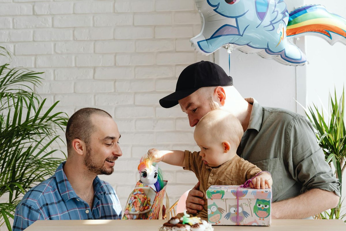 A joyful indoor birthday celebration with same-sex parents and their child surrounded by gifts and balloons.