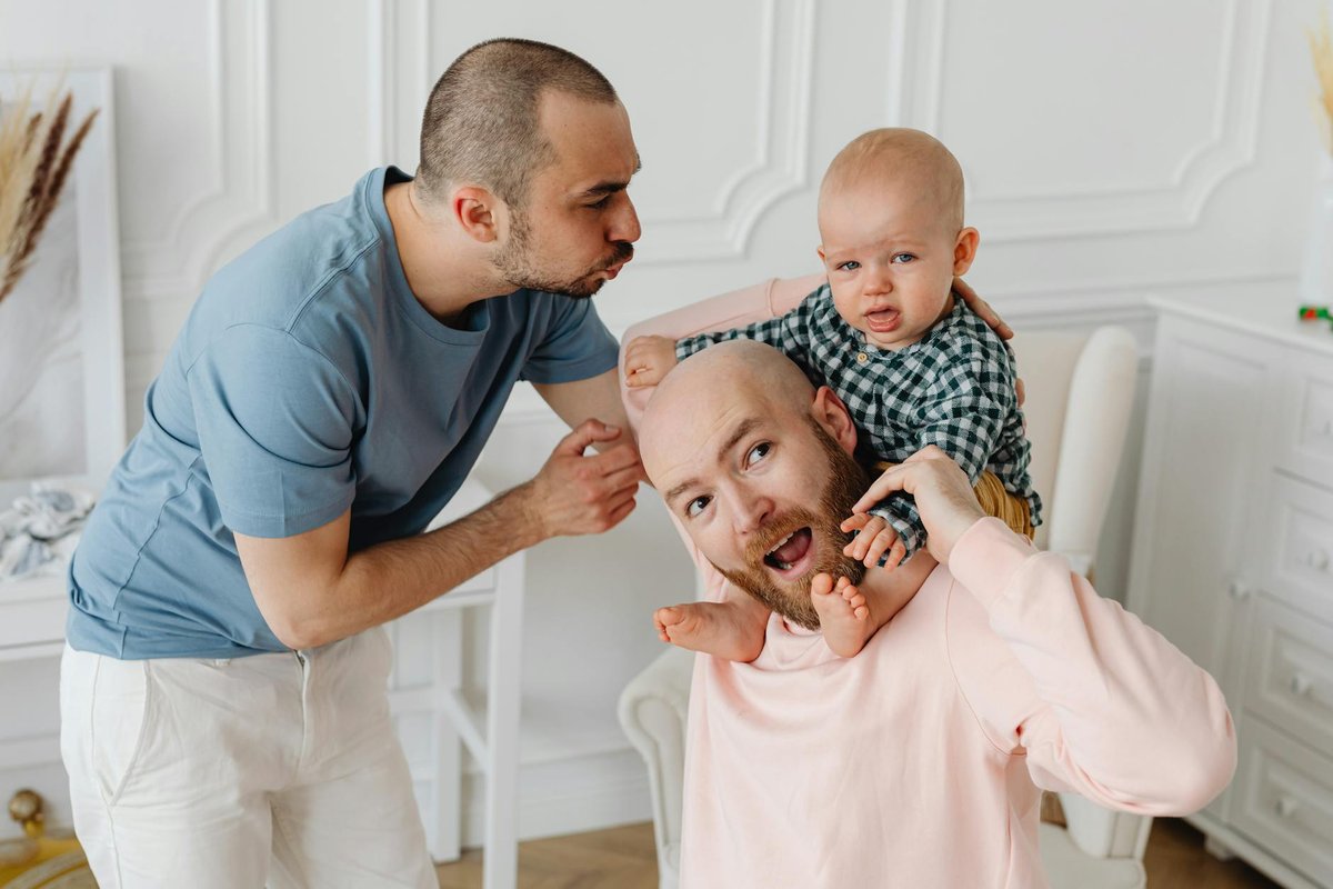 A joyful same-sex couple playing with their baby indoors, showcasing love and family bonding.