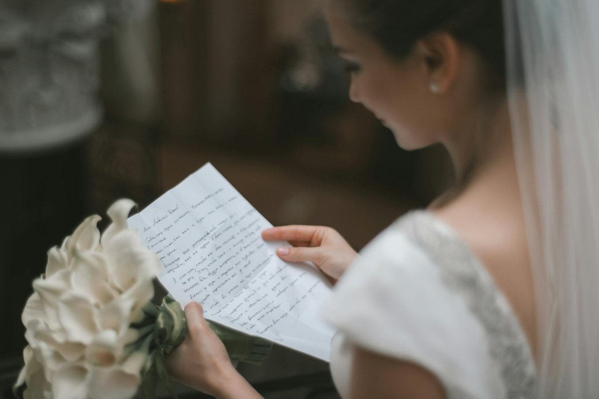 Back view of crop charming bride in white dress and veil reading wedding vow on paper sheet with bouquet of arum lilies during weeding ceremony
