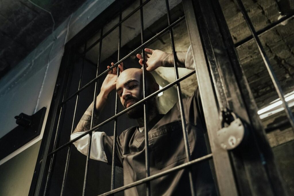 A bald prisoner with tattoos stands in a prison cell, gripping the bars.