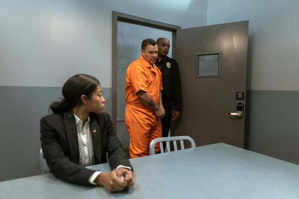 A handcuffed prisoner is escorted by a police officer into an interrogation room with a lawyer present.