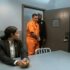 A handcuffed prisoner is escorted by a police officer into an interrogation room with a lawyer present.