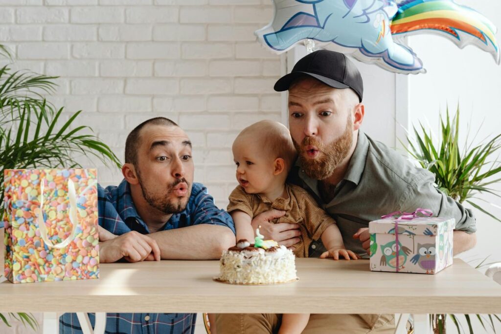 Two fathers celebrate their baby's birthday with a cake and gifts.