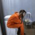 A distressed woman in an orange jumpsuit sitting in a prison cell, head in hands.